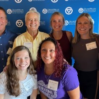 Group of 7 GVSU Alumni smiling in front of GVSU Alumni Association sign